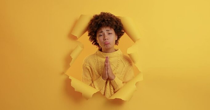 Sad dark skinned woman makes imploring expression keeps palms pressed together asks for favor says please begs permission wears casual jumper poses through torn yellow background. Let me do this