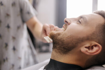Obraz na płótnie Canvas Professional hairdresser uses a hair clipper for fringing beard for a handsome man in barbershop. side view