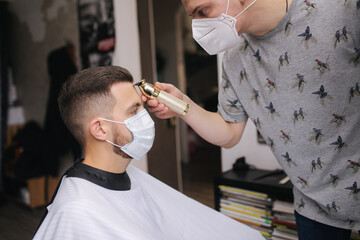 Professional hairdresser uses a hair clipper for fringing hair for a handsome bearder man. Client with face mask protect from the coronavirus. The hairdresser also wears a mask