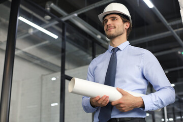 Confident young business man in hardhat holding blueprint and looking away while standing indoors.