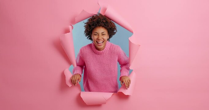 Carefree ethnic young woman giggles positively has fun and laughs at something funny dressed in knitted sweater poses in ripped hole through paper pink background. Happiness and joy concept.