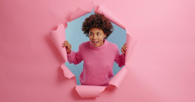 Happy smiling woman looks left and right sees something surprising dressed in knitted sweater poses through hole of pink background. Ethnic female gazes with wonder at different sides stands in studio