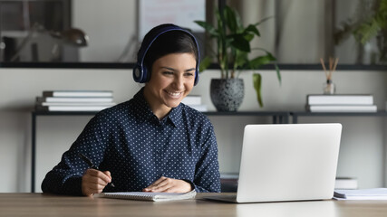 Smiling Indian businesswoman intern wearing headphones watching webinar, writing notes, listening...