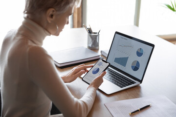 Rear view confident businesswoman holding smartphone, working with financial statistics, presentation with charts diagrams on phone and laptop screens, female employee accountant analyzing data