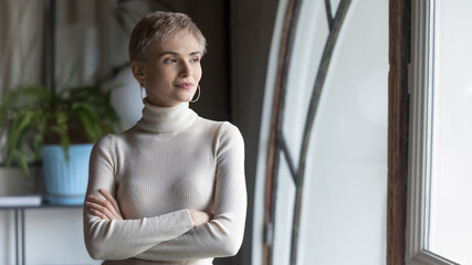 Close up confident businesswoman looking to aside through window, standing in modern office with arms crossed, thinking about future, pondering project strategy or startup idea, business vision