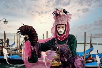 Woman in mask wearing ornate colorful carnival costume on San Marco lagoon background at sunset. ...