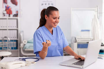 Medical nurse smiling while using laptop in hospital office wearing blue uniform. Health care practitioner sitting at desk using computer in modern clinic looking at monitor, medicine.