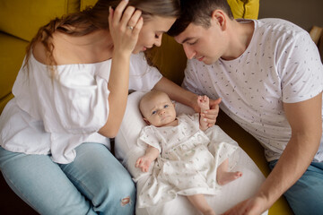 Young caucasian beautiful parents mom and dad hug and kiss their newborn daughter sitting on the sofa at home
