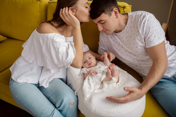 Obraz na płótnie Canvas Young caucasian beautiful parents mom and dad hug and kiss their newborn daughter sitting on the sofa at home 