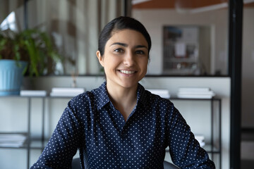 Head shot portrait smiling Indian businesswoman looking at camera, sitting in office, making video...