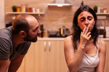 Woman suffering after husband beating and smoking sitting on chair in kitchen. Violent aggressive man abusing injuring terrified helpless, vulnerable, afraid, and panicked woman.