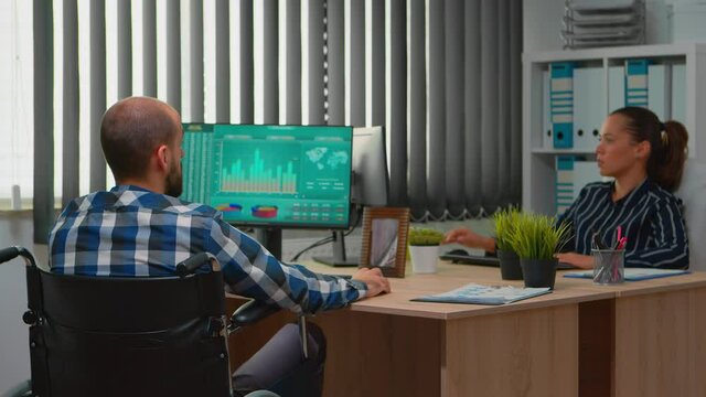 Disabled entrepreneur sitting in wheelchair immobilized analysing financial economy statistics on computer in business office discussing with colleague. Handicapped businessman using modern technology