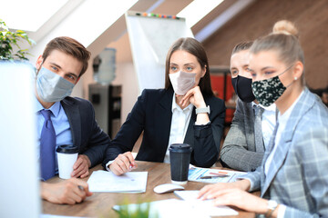 Group of young business people working, communicating while sitting at the office desk together with colleagues in preventive masks during epidemy.