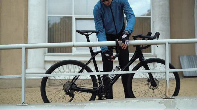 a male cyclist approaches his bike, removes the combination lock, sits down and starts to ride