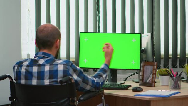 Immobilized businessman in wheelchair using computer with chroma key for videomeeting. Handicapped disabled freelancer looking at pc with green screen, mockup, key talking with remotely colleagues