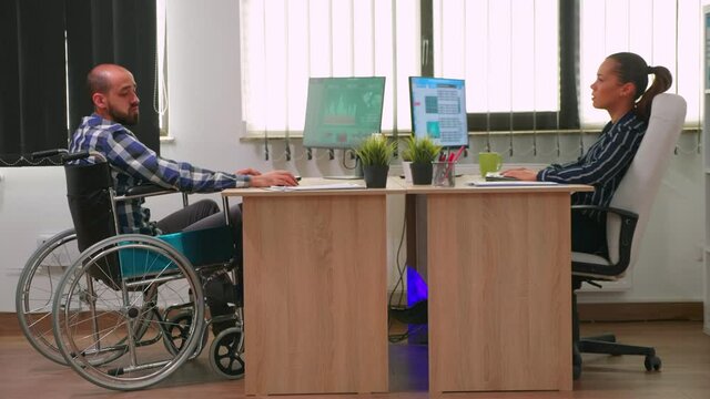 Businesswoman talking with paralysed colleague analyzing financial statistics, looking at desktop and typing. Disabled, immobilized freelancer working in corporate company using modern technology.
