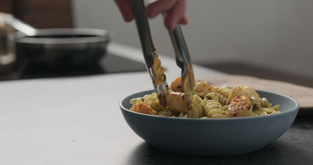 Slow motion add scallops to pesto fusilli in blue bowl on concrete countertop