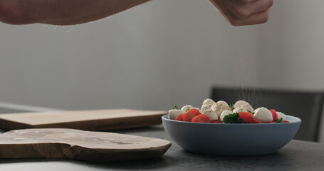 Slow motion man seasoning salad with kale, tomatoes and mozzarella in blue bowl on kitchen countertop