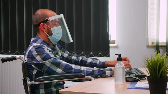 Disabled entrepreneur sitting in wheelchair with protection mask cleaning hands before typing on computer in business modern office. Handicapped freelancer with visor respecting social distance