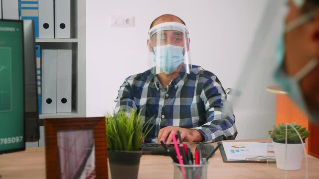 Handicapped businessman with visor and protection mask discussing with colleague working in new normal business office. Man respecting social distance in financial company during global pandemic.