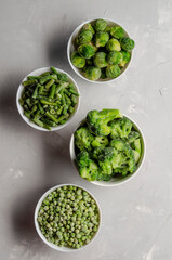 A mix of frozen green vegetables: beans peas broccoli Brussels sprouts in bowls on a grey background. Top view
