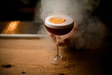Close-up view on glass of drink with foam stands on smoky bar counter.