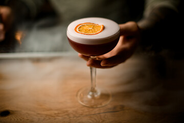 man's hand holding glass of drink with foam at smoky bar counter.