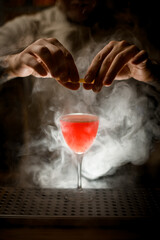 Close-up view of bartender holding lemon peel and sprinkles on glass with cocktail at smoky bar.