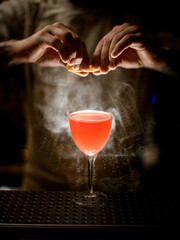 man bartender holding lemon peel and sprinkles on glass with cocktail at smoky bar.