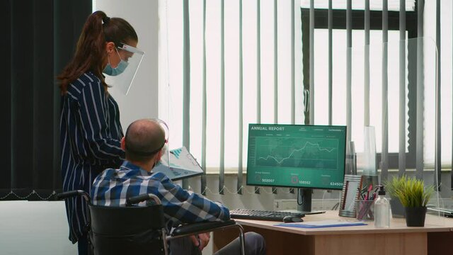 Woman pointing on desktop talking with disabled colleague in new normal office in front of computer. Team of financial expert working on computer analysing economic graphs respecting social distance.
