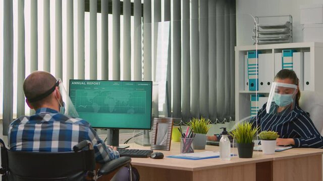 Businesswoman analysing financial statistics talking with handicapped coworker sitting in wheelchair working in new normal business office wearing face protection against covid-19.