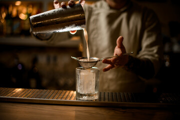 bartender holds sieve over old fashioned glass and gently pours cocktail from shaker.