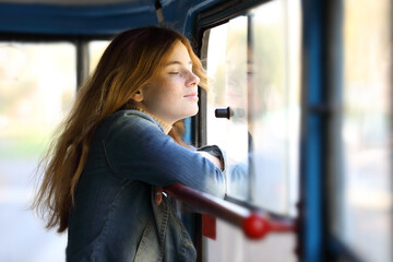 Young pretty red-haired teenage girl standing by the window of a moving tram - obrazy, fototapety, plakaty
