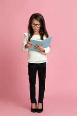 fashionable cute little child girl in oversized mother's shoes holding notebook on pink background. child playing businesswoman or teacher