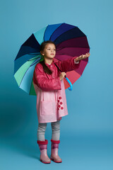 little child girl with multicolored umbrella in pink rain coat and rubber boots on blue background. girl checking for rain