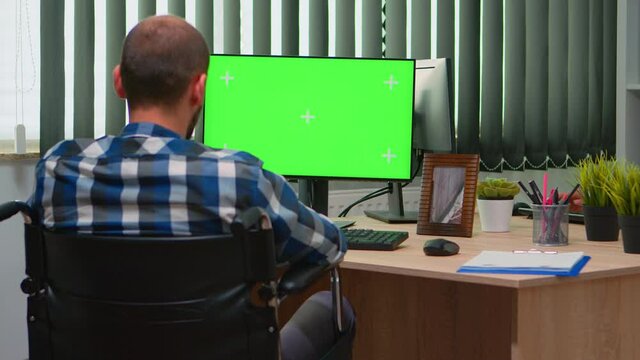 Handicapped disabled freelancer looking at pc with green screen talking with colleagues. Immobilized businessman in wheelchair using computer with chroma key, mockup, greenscreen for videomeeting.