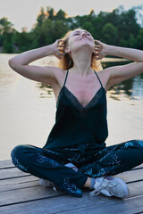 Beautiful blond young woman in dark green lace top pajamas pants sitting near the river bank with forest reflection in the water on blur background 