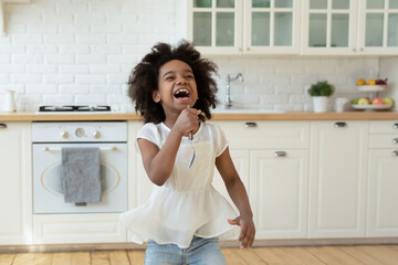 Overjoyed little African American girl holding fork as microphone and singing, standing in modern...