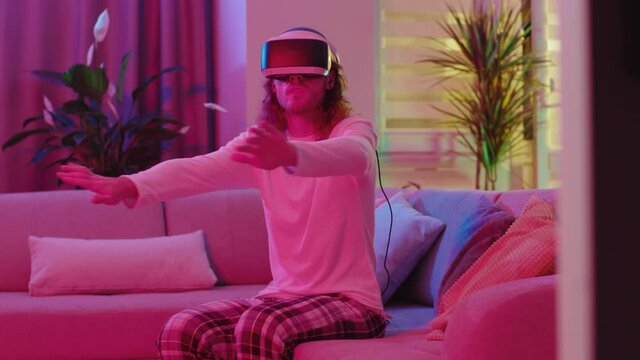 Energetic gaming man young hipster adult wearing VR headset playing online virtual 3d simulation diving swimming game sitting on sofa staying home.