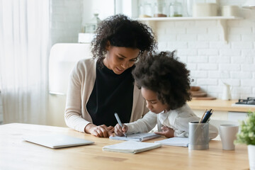 Smiling caring African American mother helping to adorable little daughter with homework, sitting at table in modern kitchen, pretty girl doing school tasks, studying, writing with pen, homeschooling