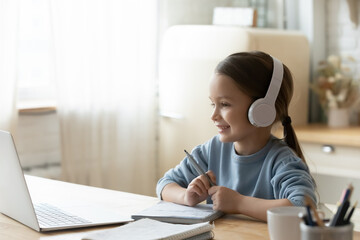 Smiling little girl wearing headphones using laptop, studying online, looking at computer screen,...