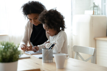 Caring African American mother helping to little daughter with homework, doing school tasks...