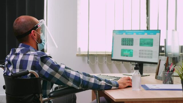 Disabled businessman sitting in wheelchair wearing face mask and visor against coronavirus disinfecting hands before checking financial data in business office. Freelancer respecting social distance