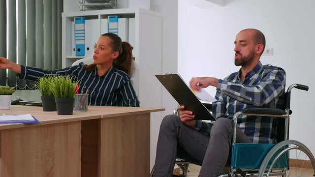 Handicapped company manager in wheelchair giving advice about financial project to partner in office room company room analysing graphs from documents. Discussing and pointing on computer desktop