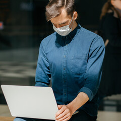 young man in a protective mask is working on a laptop, sitting on a bench.