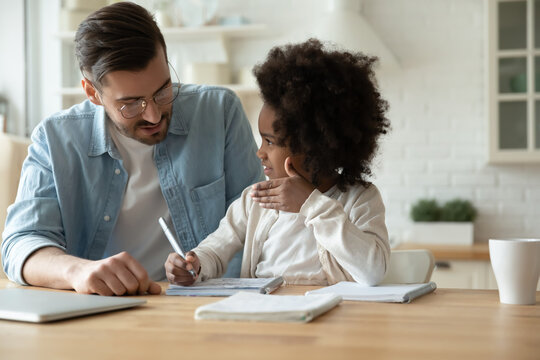 Caring father wearing glasses helping to adorable little daughter, pretty African American child girl with dad studying, doing school homework, writing, multiracial family, homeschooling concept