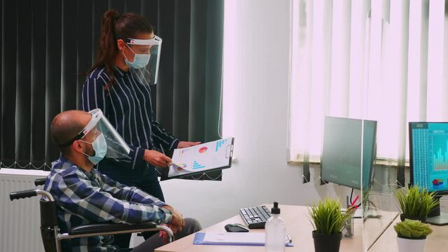 Business people wearing protective mask in new normal office discussing strategy, businessman sitting in wheelchair. Team working in profesional workspace in corporate company during covid-19 pandemic