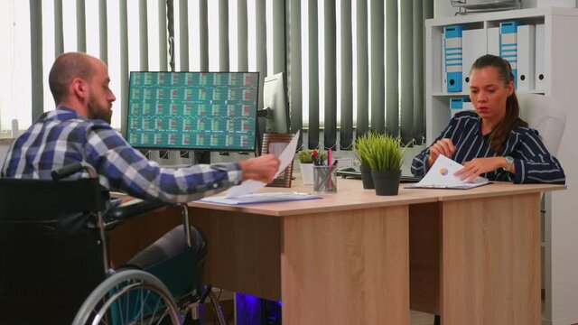Businessman in wheelchair working in corporate office room changing documents analysing graphs. Employees in modern company typing on computer keyboard and pointing at desktop checking clients list.