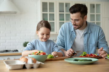 Caring father and little daughter chopping fresh vegetables on board, cooking salad together, sitting at wooden table in kitchen at home, preparing meal, family enjoying leisure time