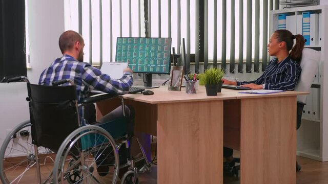 Paralysed financial expert sitting in wheelchair analysing economy statistics of company in business office discussing with woman manager. Handicapped, immobilized businessman using modern technology.